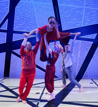 Three dancers lift one dancer up in the air on stage in front of a blue background.