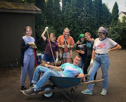 Image shows a group of students with tools for working outdoors. 6 students are standing and one is lounging in a wheelbarrow.