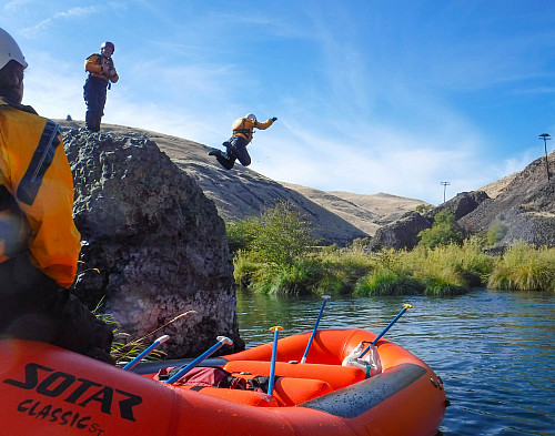 Image shows a tall boulder on the right side of the image sticking out of a river. There is one person standing on top of the boulder and...