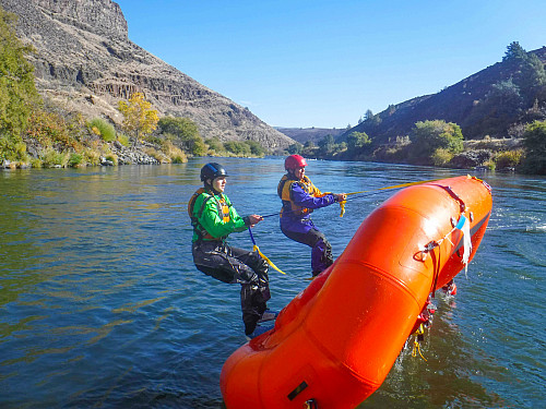 Image shows two people wearing drysuits, PFD's, and helmets. They are standing on the bottom of a flipped orange raft holding onto straps...