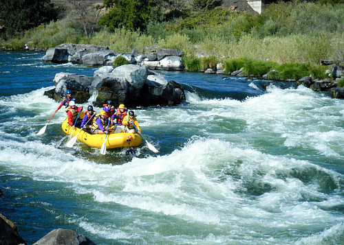 Image shows a yellow raft on the left side of the image entering a rapid on the white waters of the Deschutes River. The raft is full of ...