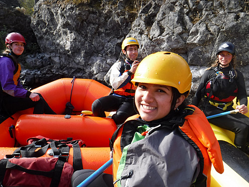 Image shows a person in a helmet and PFD sitting in an orange raft and smiling at the camera. Behind them are three other people also smi...