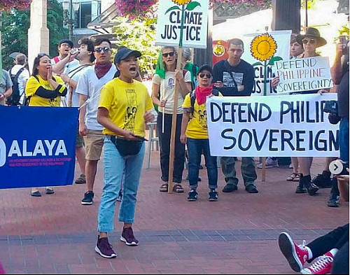 Image shows a group of people marching. In the front is a woman in a yellow shirt and and bluejeans talking to the crowd. Signs read Defe...