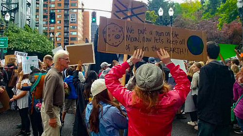 Image shows people at a Climate March in Portland. In the foreground is a person in a red jacket holding a sign that reads, “We put...