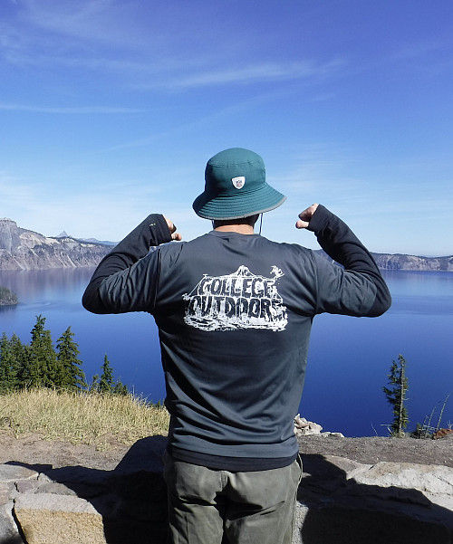 A student poses in front of a lake with a teal sun hat and two thumbs pointing to the back of a grey College Outdoors shirt.