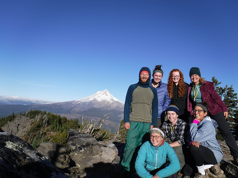 Image shows a group of people posing with a view of Mt Hood and blue skies behind them.