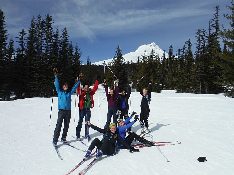 A group of participants pose in front of Mt. Hood wearing cross-country skis with their hands in the air smiling.