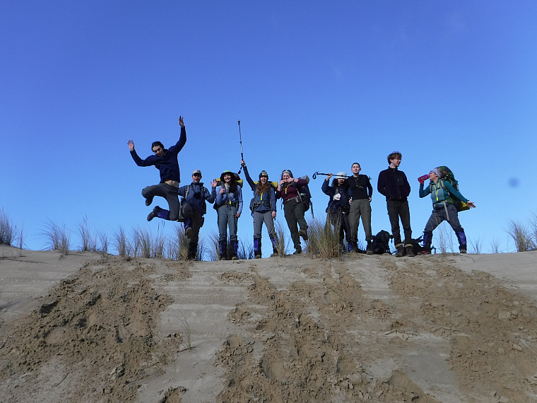 Participants pose on a sand dune with their backpacks on and hiking poles in the air smiling with blue sky behind them.