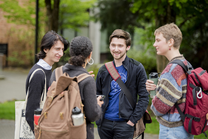 Four students, in jackets and backpacks, standing outside on campus and chatting.