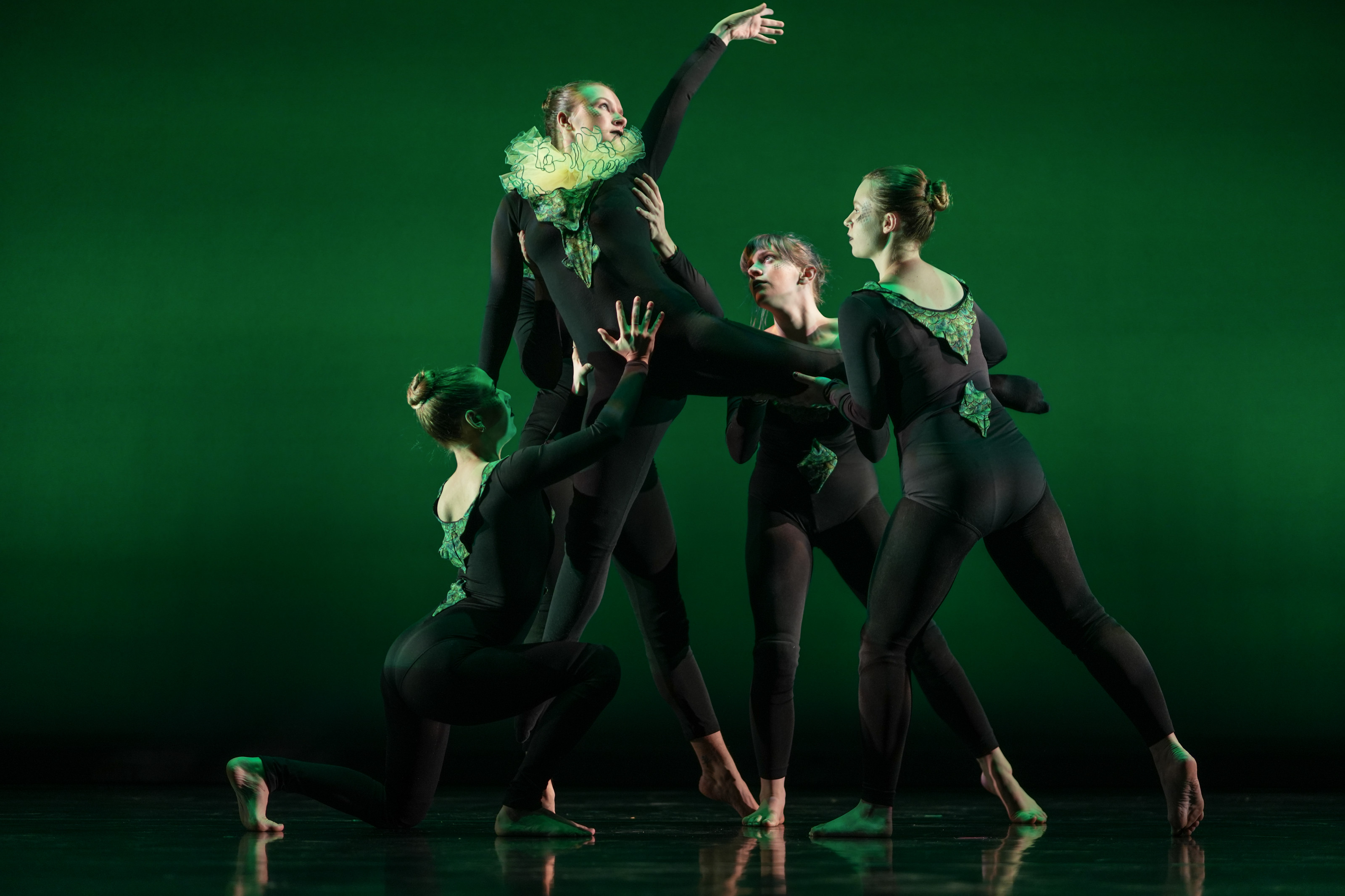 Group of students dancing against a green background. Four students lift up one student in the air.