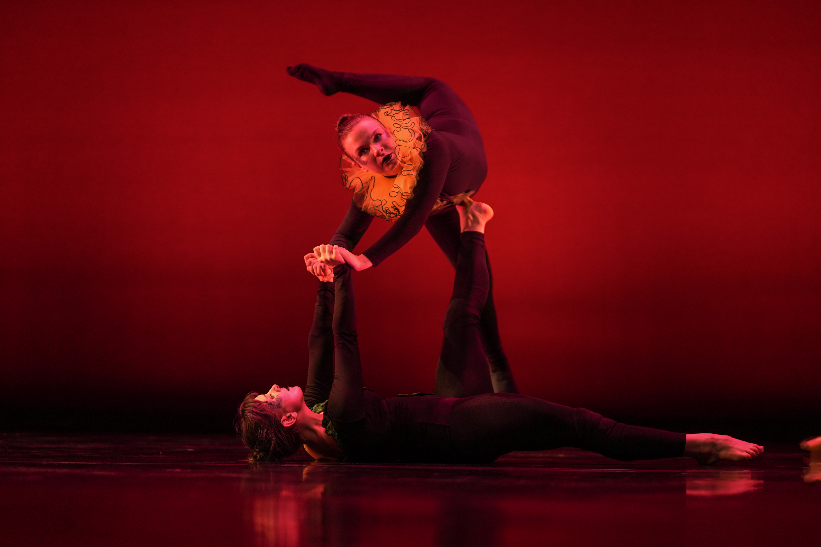 Two students against a red background. One student is on the ground lifting the other student in the air using their hands and feet.