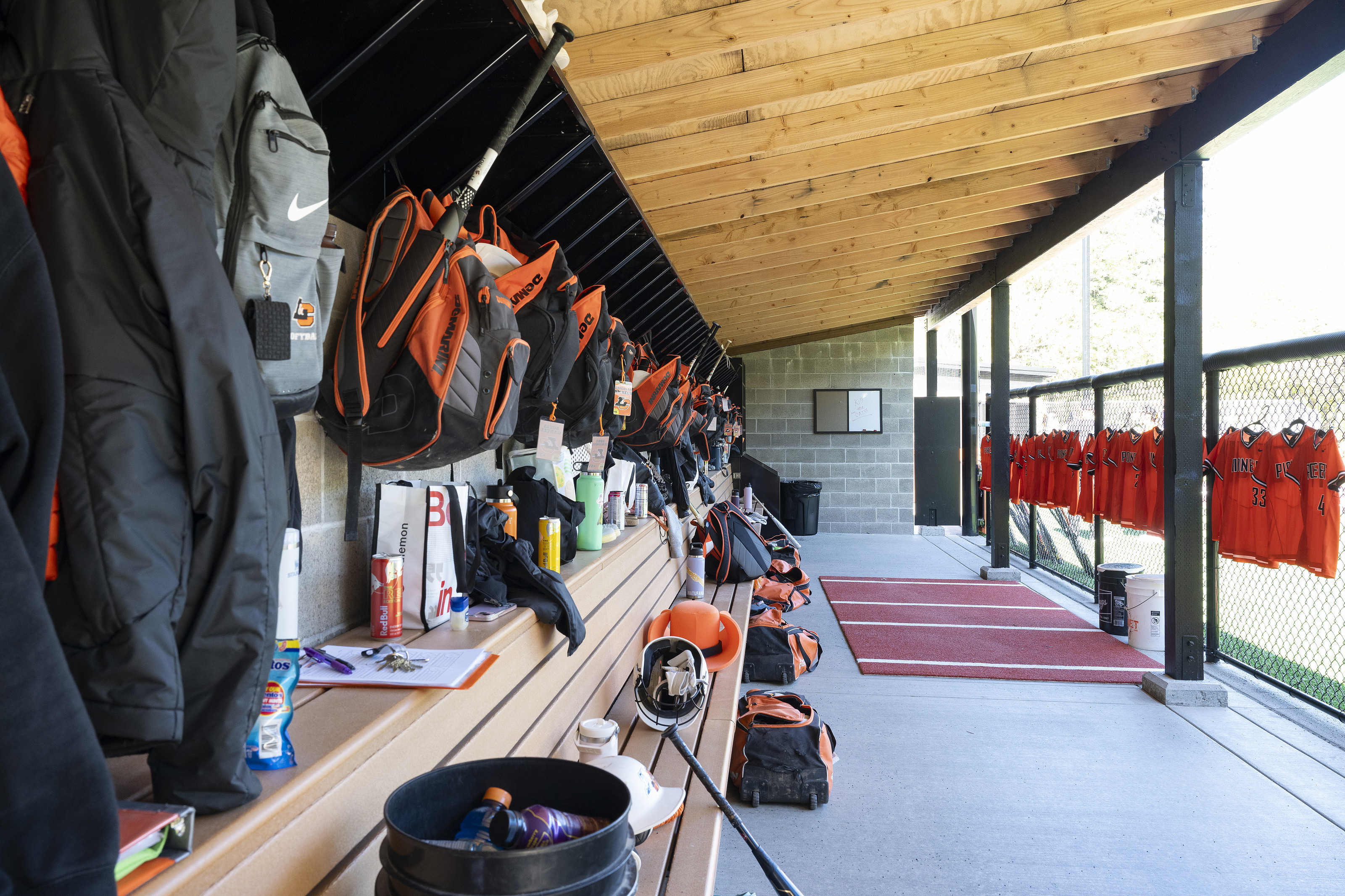 Backpacks hanging on a wall above a long wooden bench inside a softball dugout.