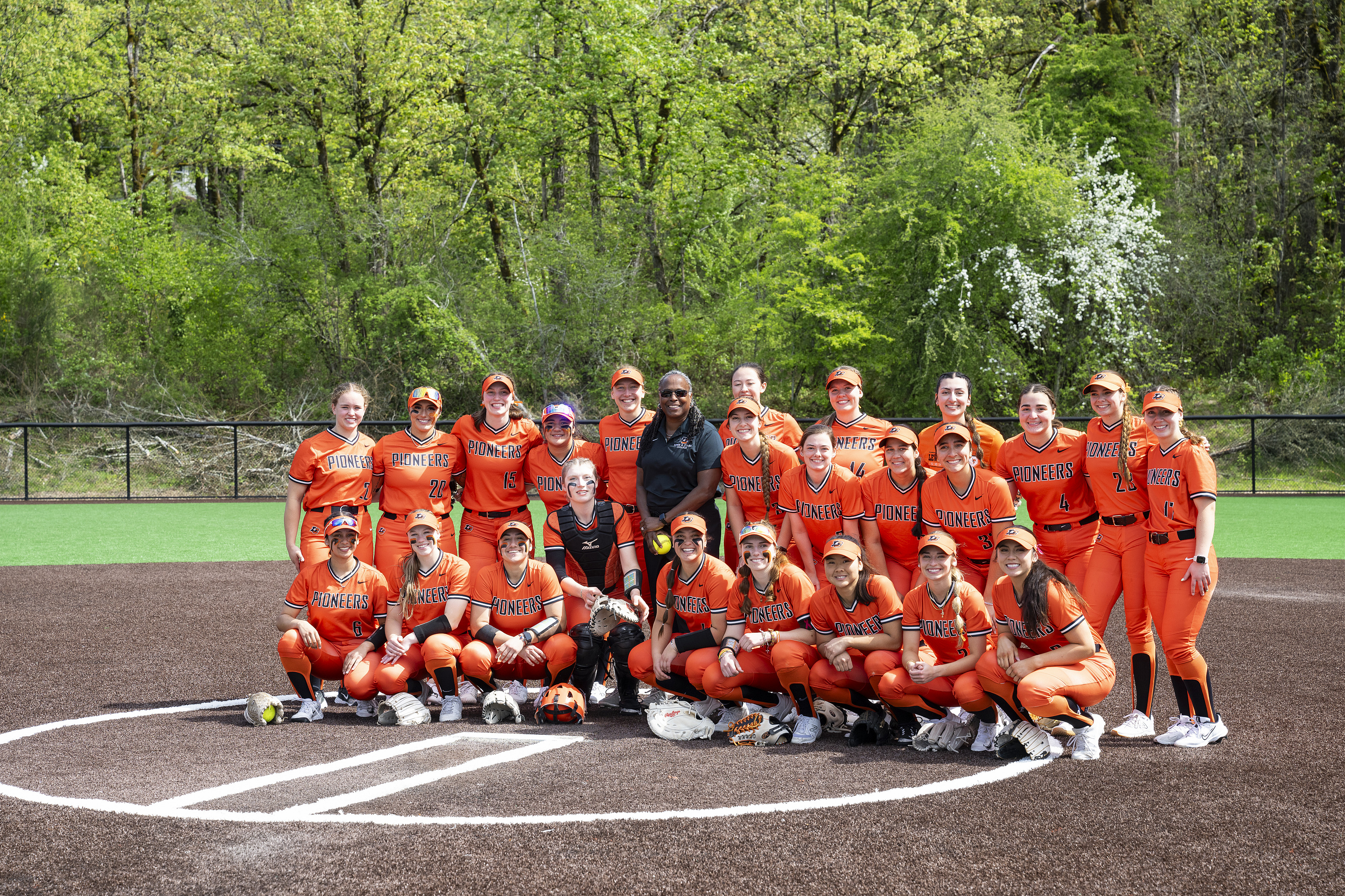 A group shot of student softball players wearing orange uniforms with President Holmes-Sullivan standing in the middle of the group.
