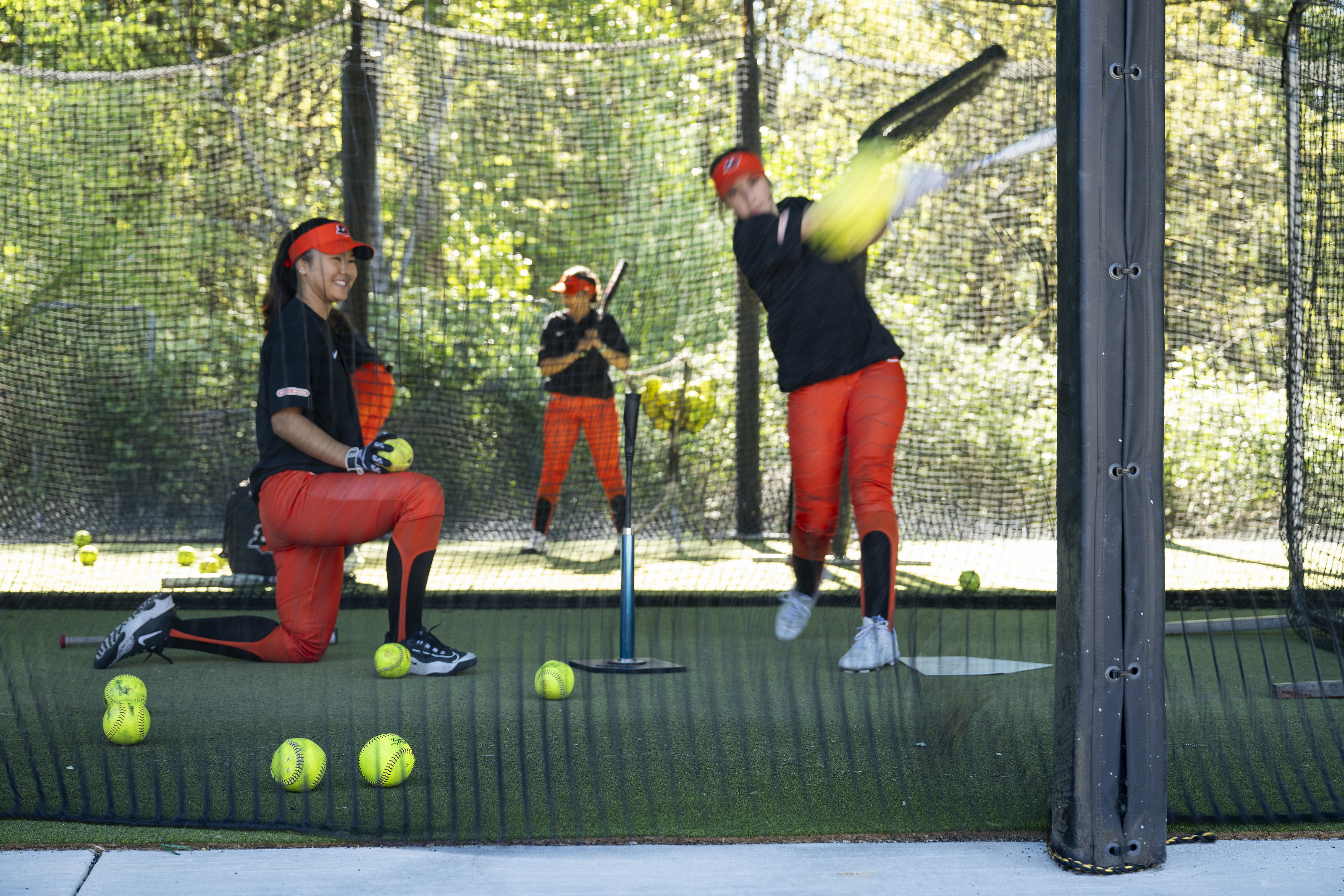 A softball player hitting a ball in a batting cage, while another player kneels nearby with a ball in her hand.