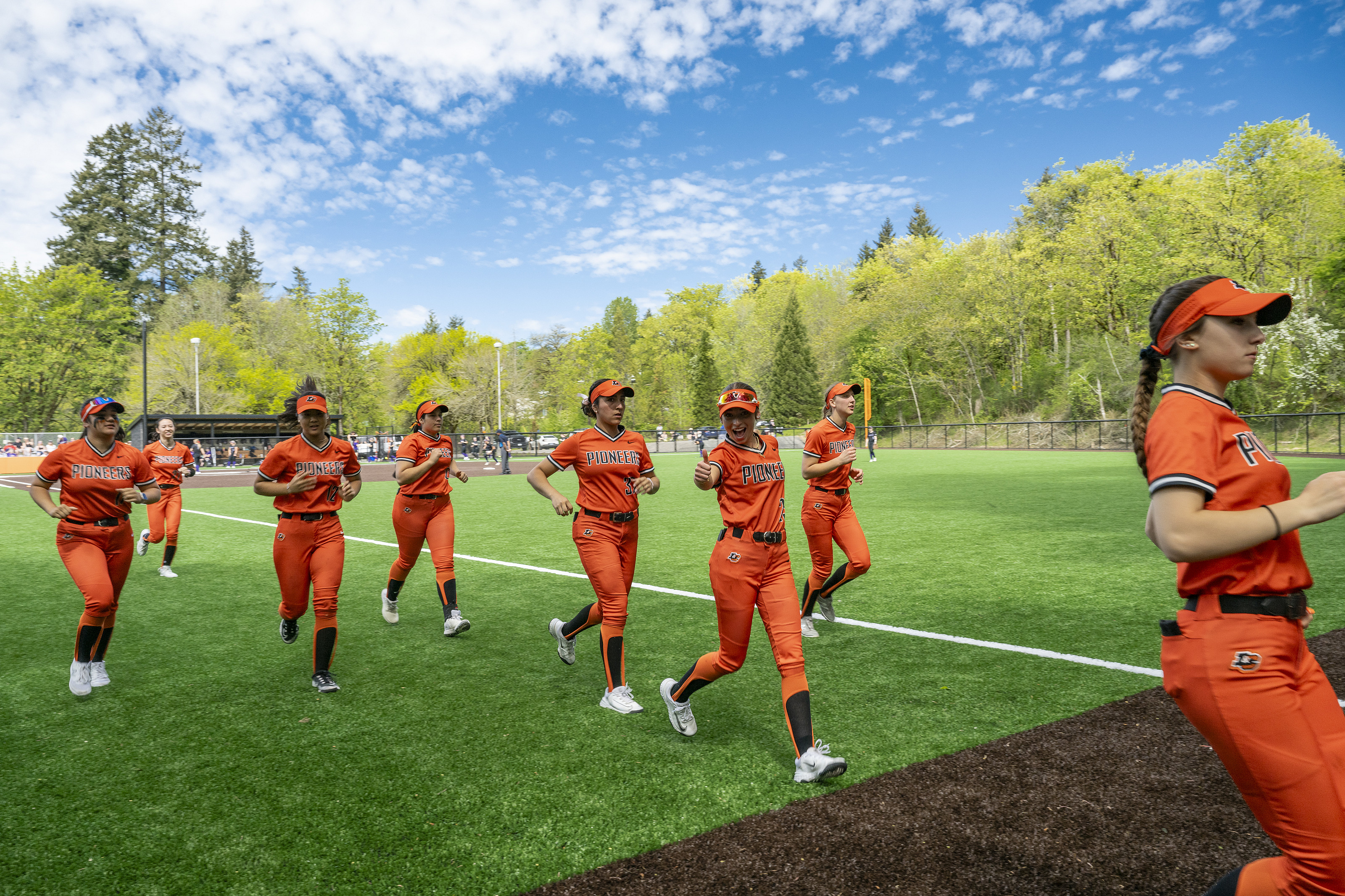 Softball players running on the field in orange uniforms. One of the players is giving a thumbs up to the camera.