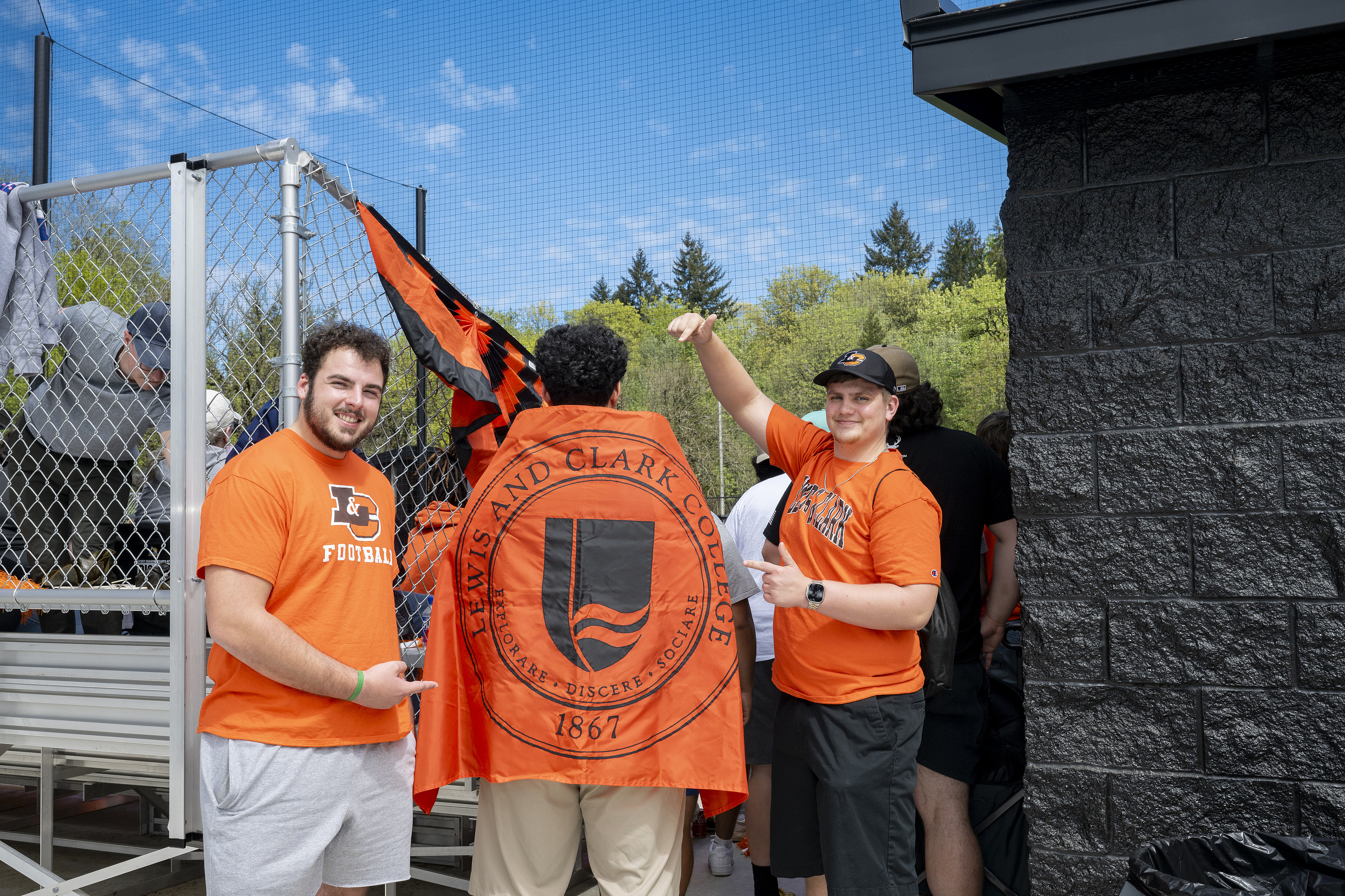 Two students wearing orange L&C athletic shirts point at a third student, with an orange L&C flag draped over his back.