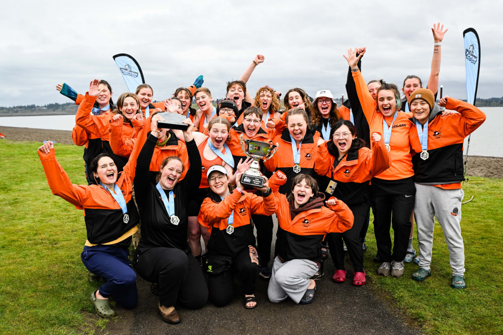 Women's rowing team wearing L&C orange jackets and cheering with their trophy.