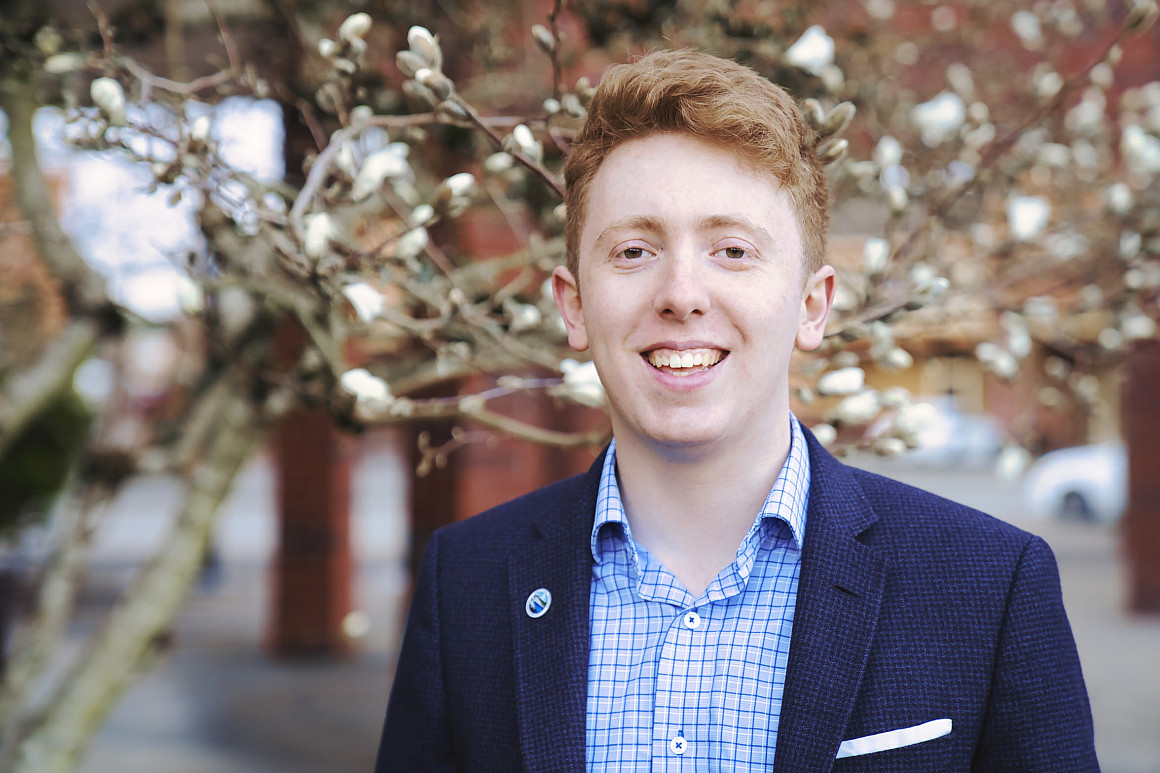 Rory posing outside wearing a blue shirt and navy suit jacket.