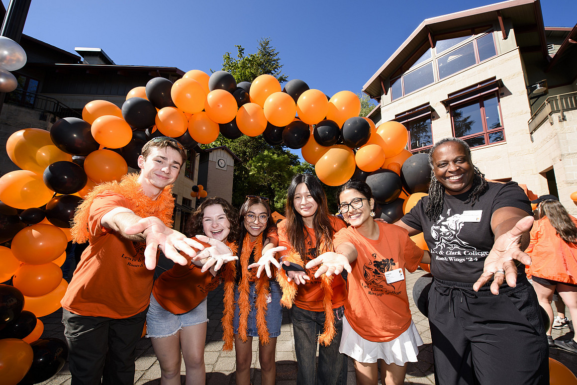 The president and a group of students smiling and reaching their hands out to the camera. A black and orange balloon arch is behind them.