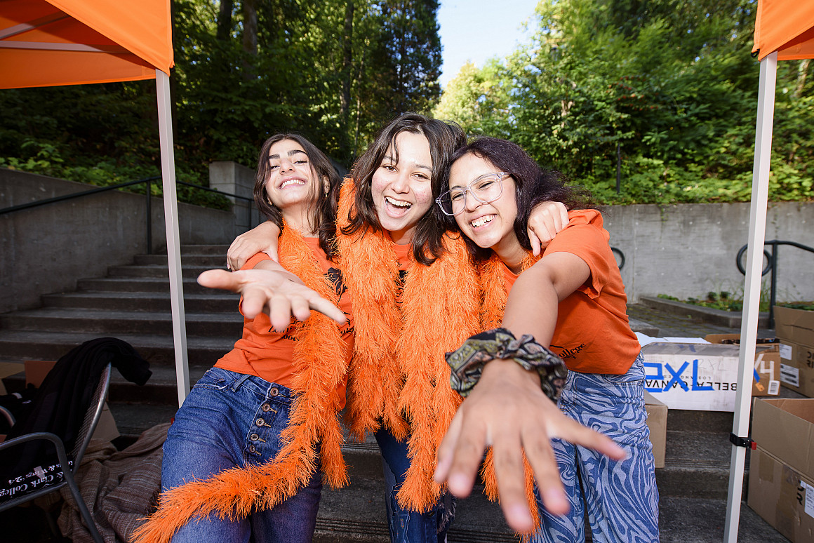 Three students wearing orange shirts and boas pose with their arms outstretched.