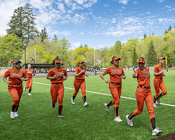 Softball players running on the field in orange uniforms. One of the players is giving a thumbs up to the camera.