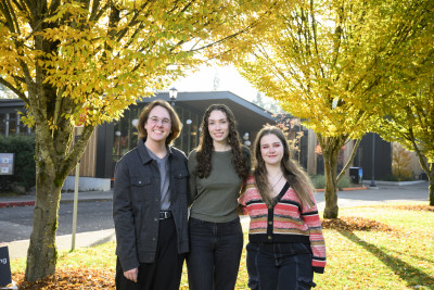 Three students smiling at the camera outdoors with lots of trees with yellow leaves surrounding them.