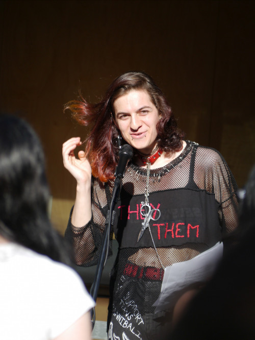 Cleo performing their piece, wearing a fishnet long-sleeve shirt over a black tank top with the words they/themin red on it.