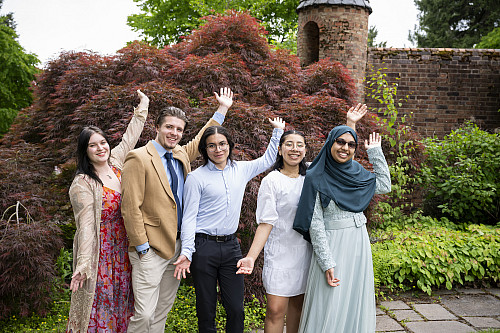 Five students posing outside, smiling and raising one arm each. They are dressed in formal clothing.