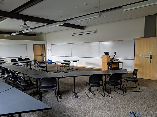 An instructor's podium is located in front of a wall of whiteboards in a classroom. The podium is surrounded by tables and chairs.