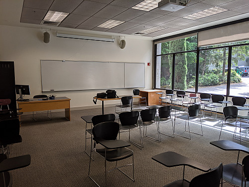 Tables and chairs fill a bright classroom. At the front of the room on the left is an instructor's table with monitor, keyboard, and mouse.