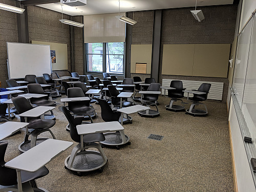 A classroom filled with chairs with attached desks. At the front of the room are two whiteboards, and a portable whitebord is in back.