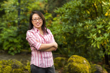 Sharon smiling outside, wearing glasses, a pink and white striped button-down shirt, and gray pants.